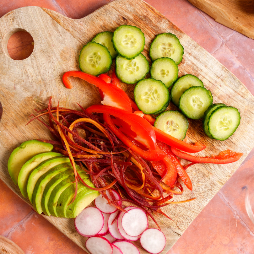 Sliced vegetables including cucumbers, red bell peppers, avocado, and radishes on a wooden cutting board.