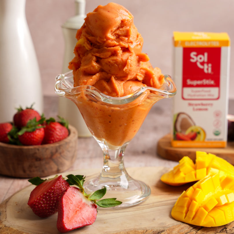 Mango sorbet in a glass bowl with strawberries and mango pieces on a wooden surface, featuring Sol-ti Strawberry Lemon SuperStix.
