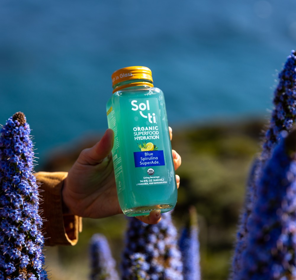 Hand holding a Blue Spirulina SuperAde organic superfood hydration bottle with a scenic background with ocean and ground