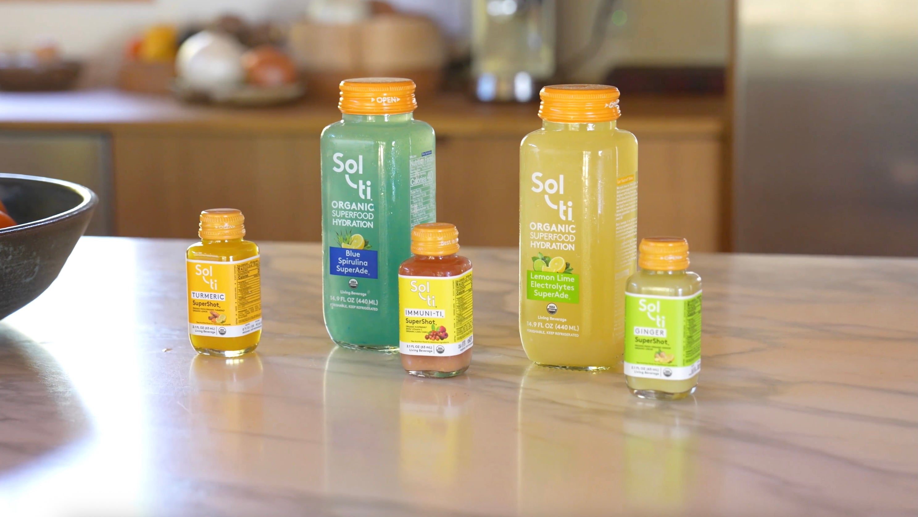 Assortment of Sol-ti organic bottled drinks on a kitchen counter, featuring two large SuperAde bottles (blue spirulina and lemon lime) and three smaller wellness shots (turmeric, IM-MUNI-TI, and ginger) with orange caps.